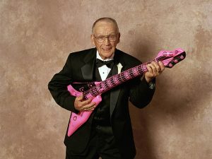 Image showing a photograph of a mature man dressed in a dinner suit holding a pink and black inflatable guitar on a page for guitar lessons